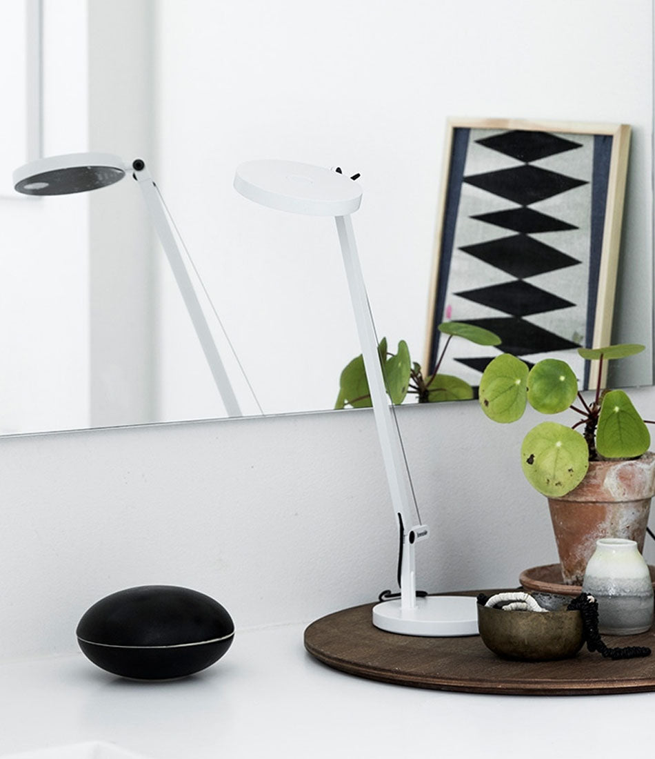White Artemide Demetra micro table lamp on a wood disc on a white counter in front of a mirror.