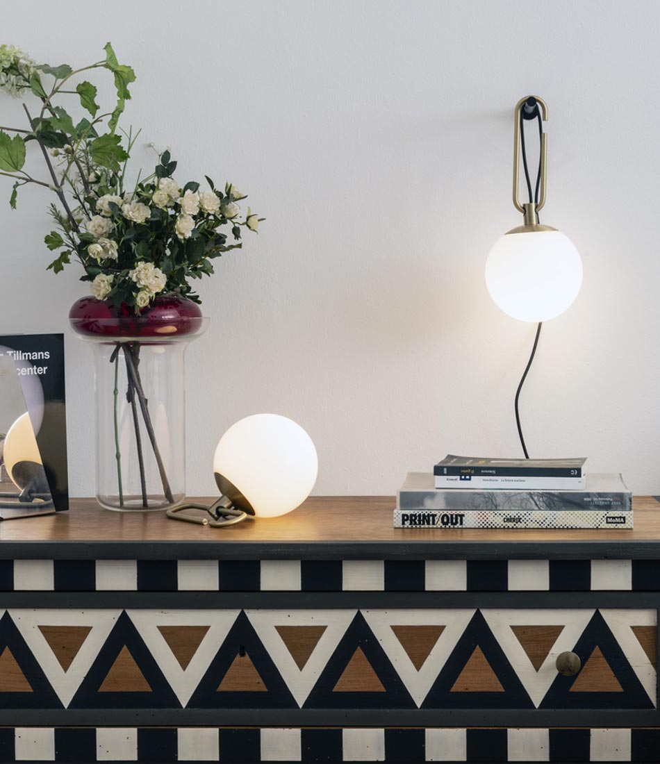An Artemide NH Wall Lamp and Table Lamp above a wooden console with books and flowers.